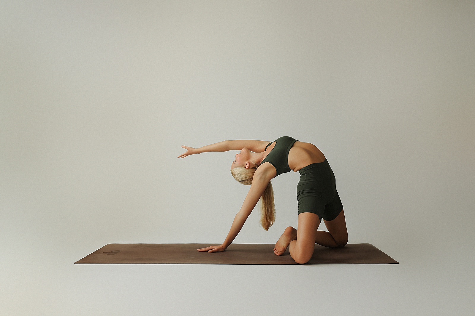 Person practicing a yoga pose on a mat, with one hand extended overhead and the other touching the mat, wearing a green outfit against a plain background.