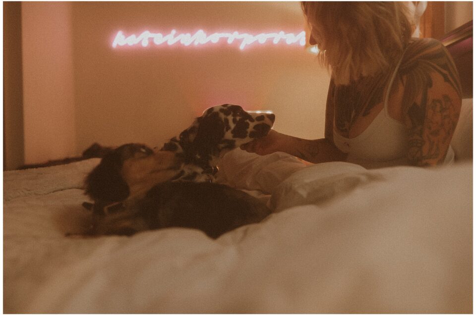 A person with tattoos sits on a bed with two dogs, under pink neon lights.