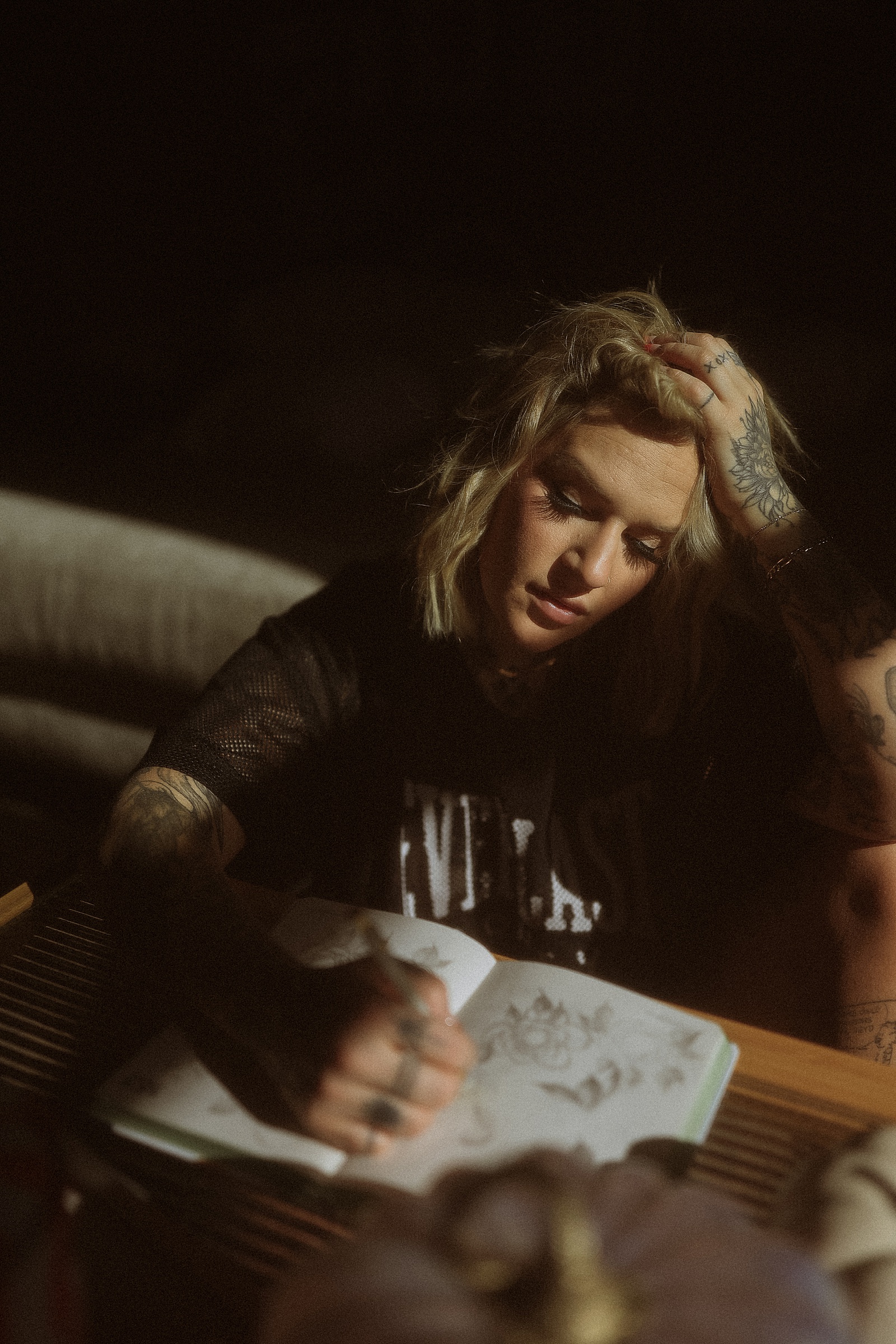 Person with tattoos, wearing a dark shirt, sits at a table, sketching in a notebook, with their head resting on one hand.