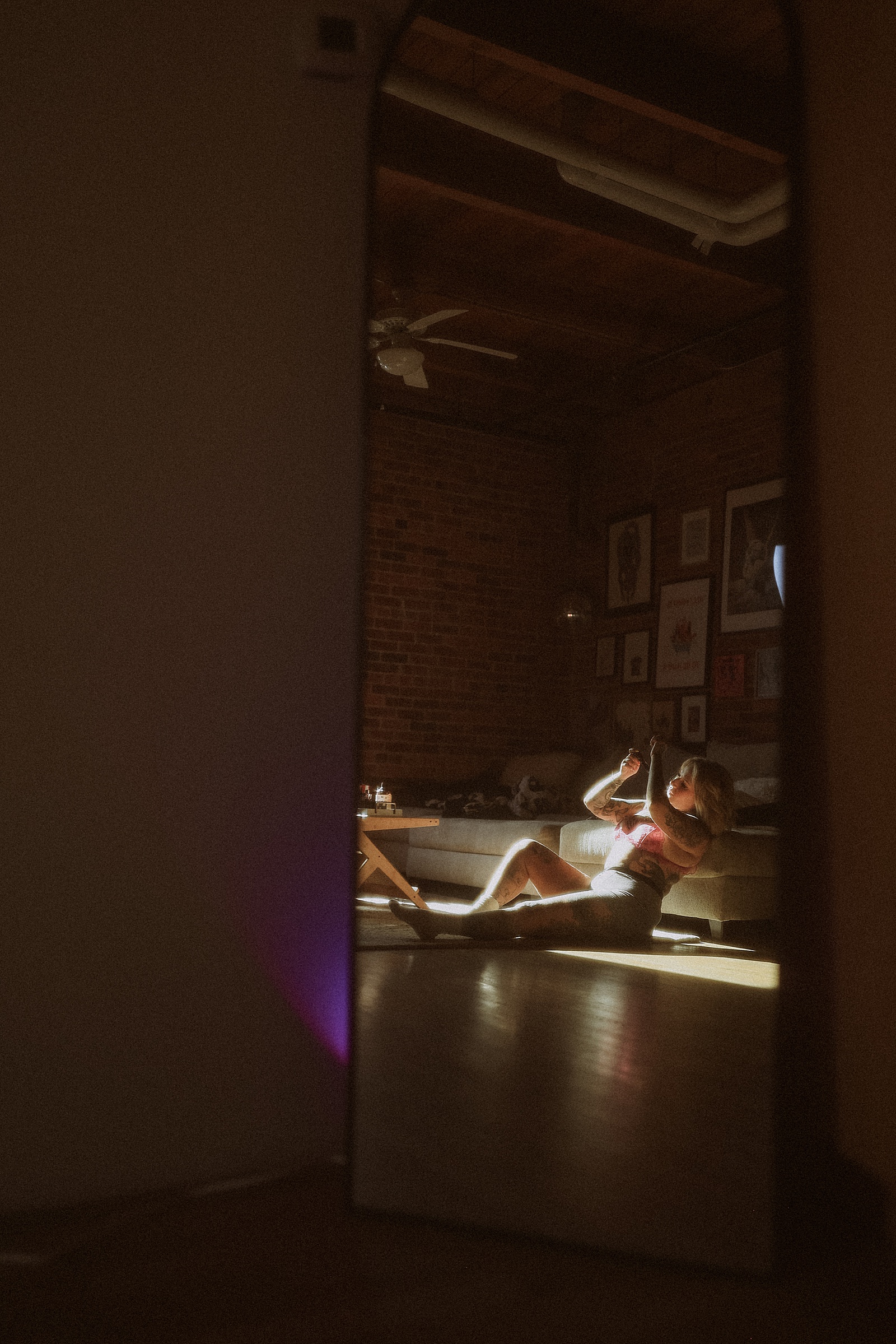 A person sits on a couch in a sunlit room, taking a photo with their phone, as seen through a large mirror reflecting the scene.