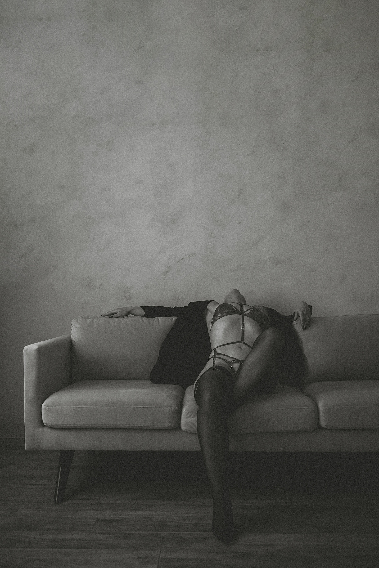 A person in lingerie and stockings reclines on a sofa with their head tilted back, in a dimly lit room with a textured wall.