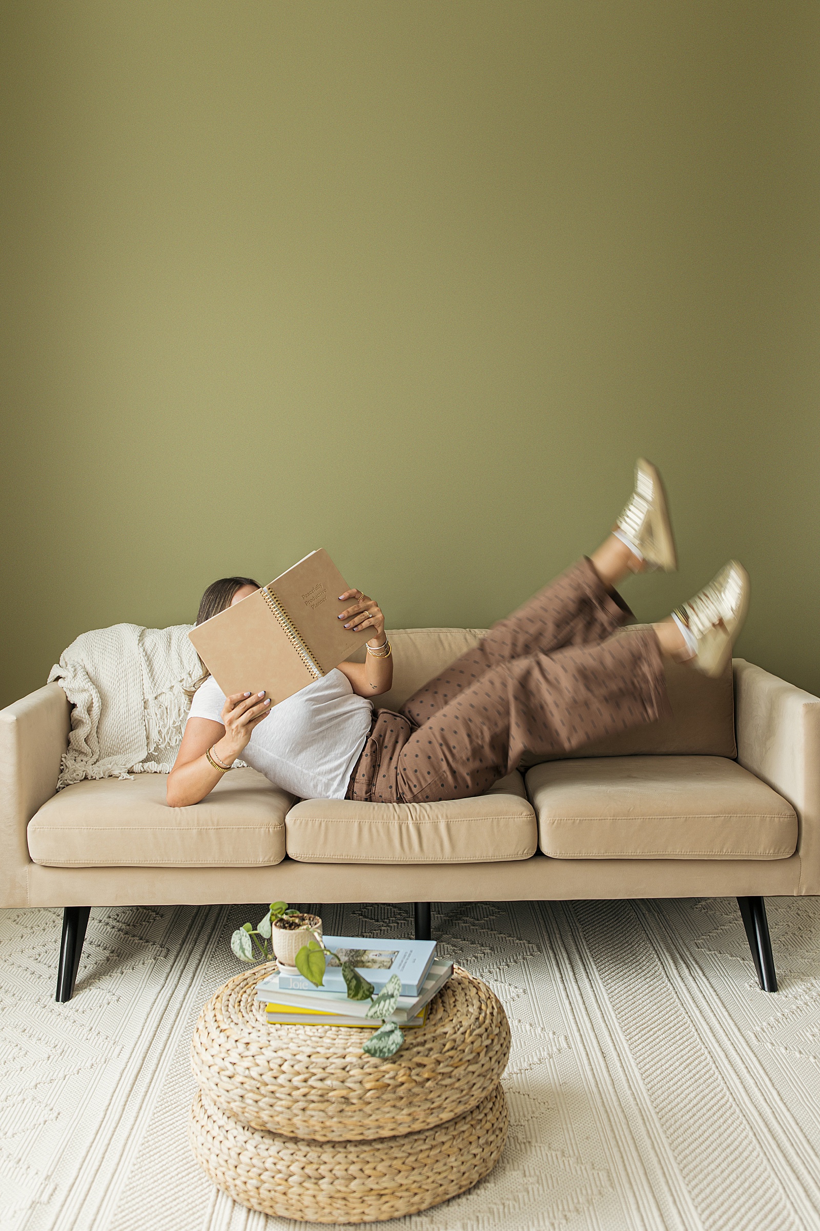 Person lying on a beige sofa with legs raised, reading a book; round woven ottoman with books and plants in the foreground, green wall background—perfect for Introverted Founders seeking calm and inspiration.