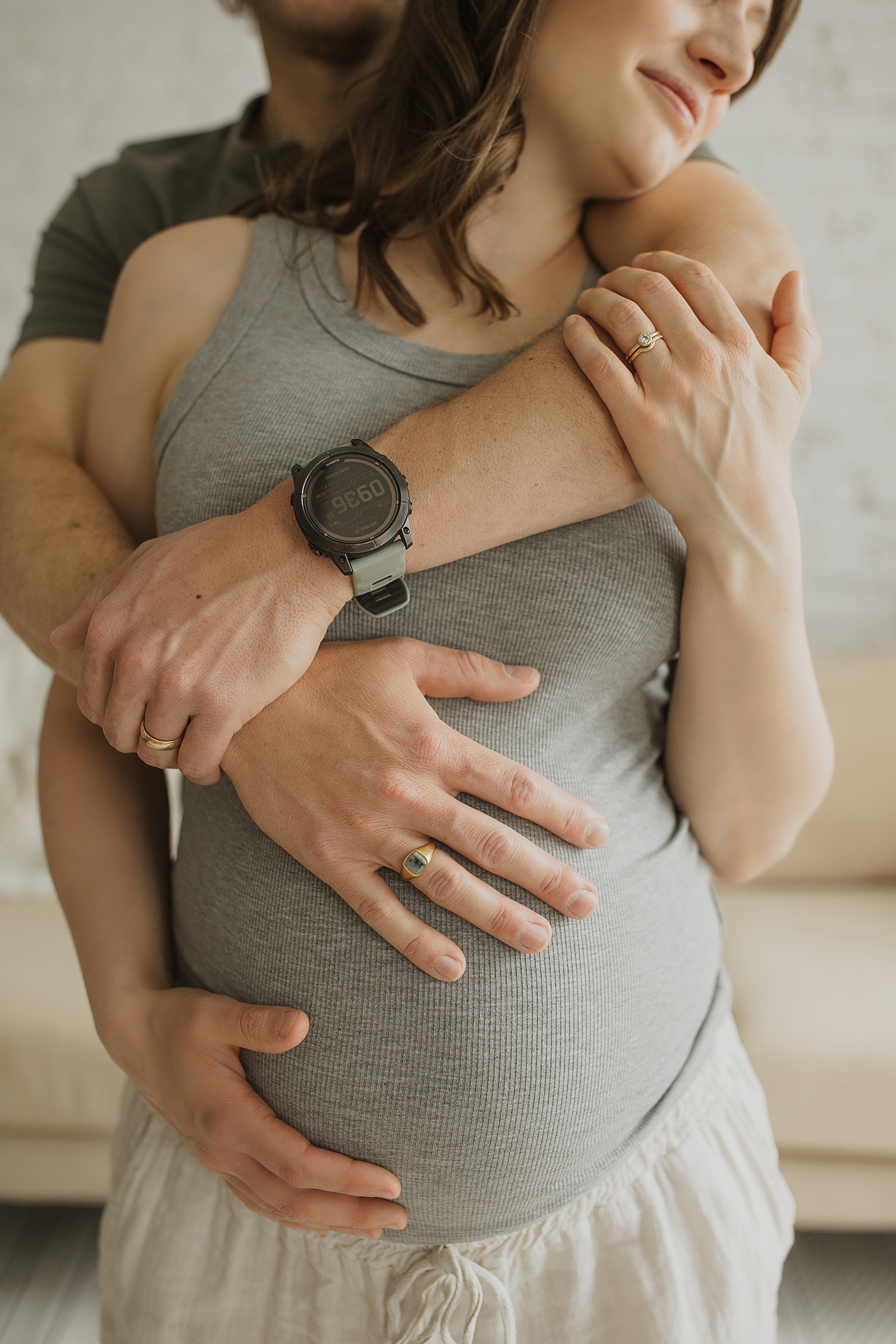 A pregnant woman in a gray tank top is embraced from behind by a man. Both have wedding rings, and his hands rest on her belly.