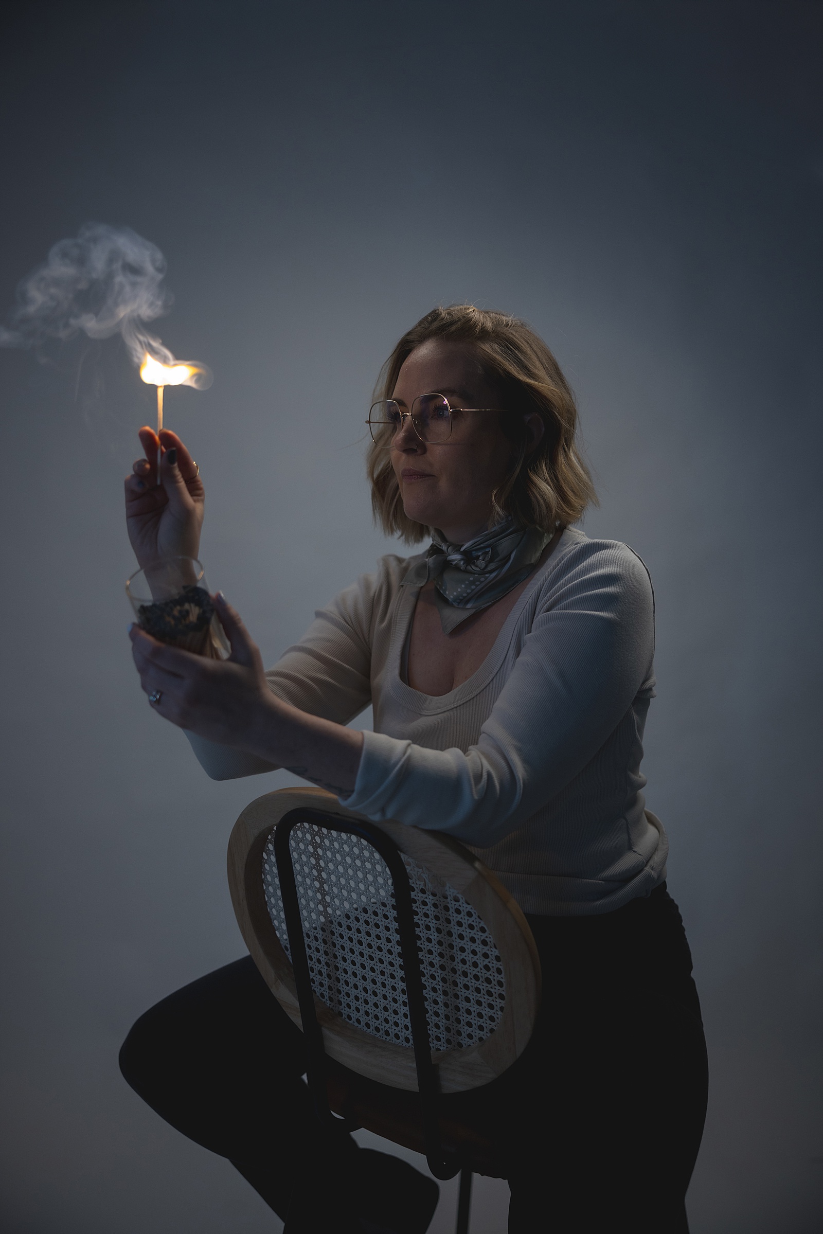 A Denver photographer for women captures a person with short blonde hair and glasses, sitting on a chair and holding a burning match above a smoking glass jar, set against a dark background.