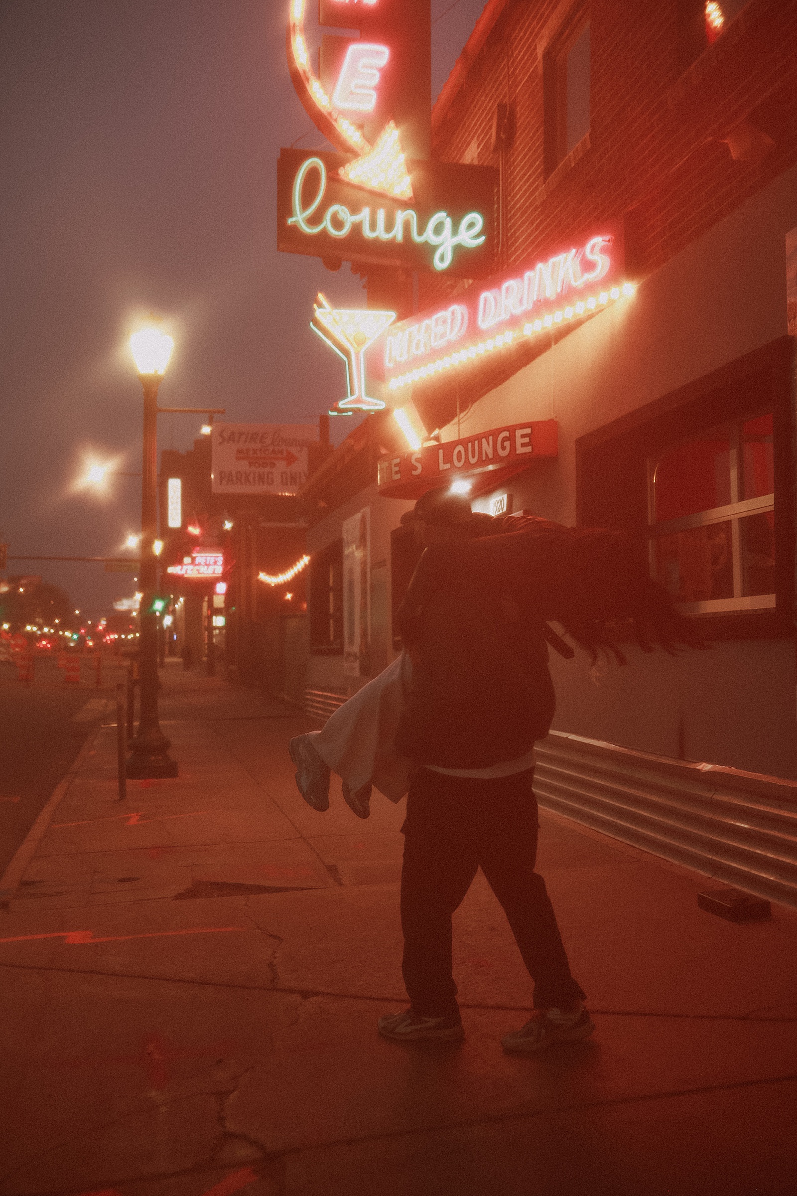 A person carries another person on a sidewalk at night near a neon-lit lounge with signs for mixed drinks, capturing the energy and romance found in cinematic couples photography.