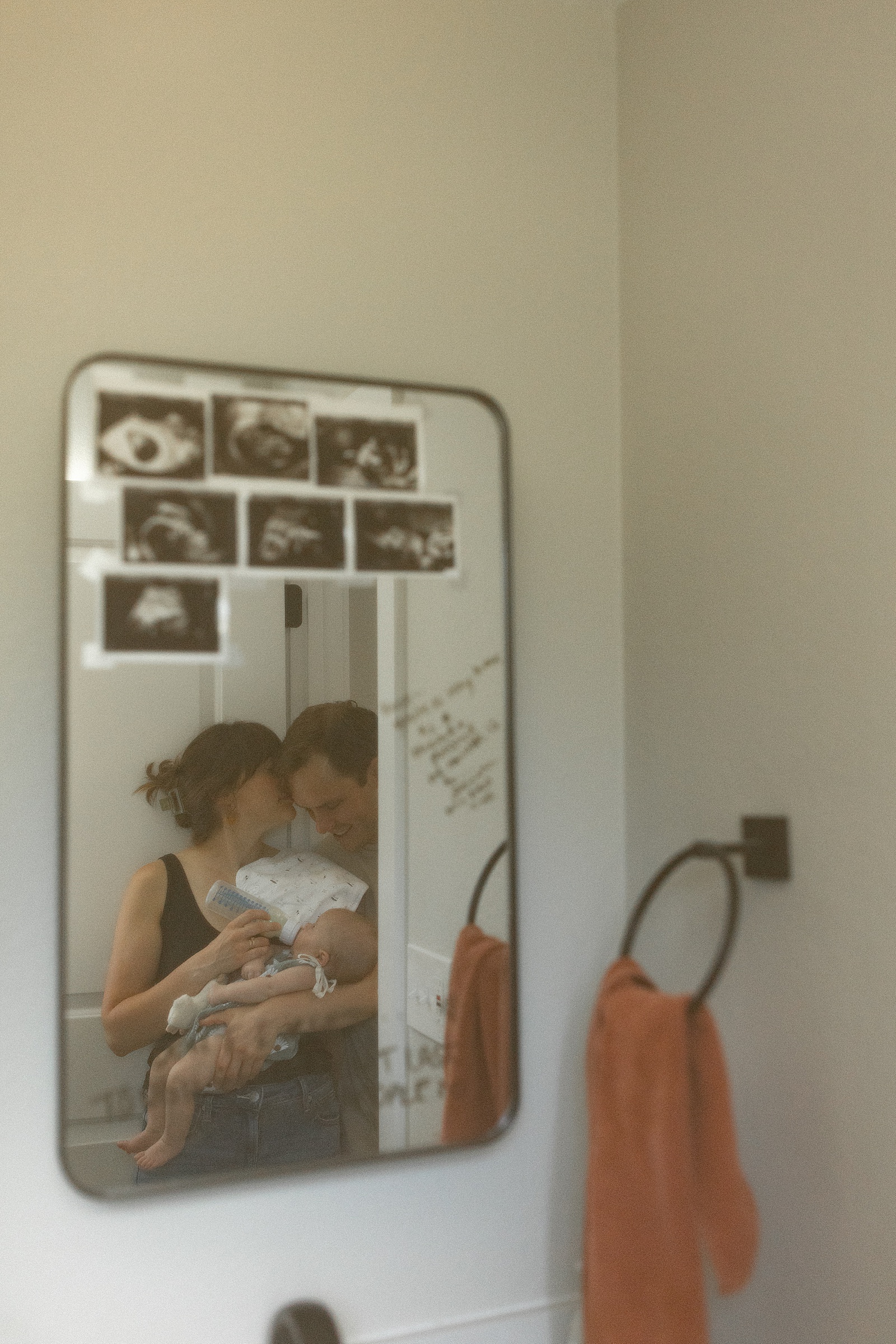 A woman and man hold a baby, reflected in a bathroom mirror with ultrasound photos and handwritten notes visible—a candid moment of in-home family photography. Two orange towels hang on the wall, adding a touch of warmth to this authentic Colorado family photography scene.