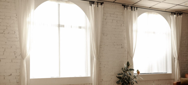 A cozy bedroom with sheer curtains diffusing sunlight through large windows, featuring an exposed brick wall and a plant in the corner.