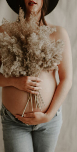 Pregnant person holding a bouquet of pampas grass in front of their belly with a neutral backdrop.