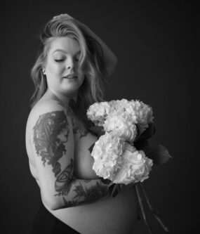 Topless pregnant woman holding flowers with hand in hair.