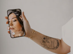 A person's tattooed arm holding up a smartphone with a Denver Makeup Artist's tutorial on the screen.