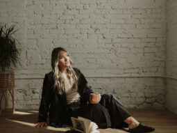 A woman in a black outfit and loafers sits on a wooden floor, leaning against a white brick wall, holding a cup and looking away from an open book.