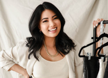 A smiling woman in a cream blazer and black leather pants stands by a clothing rack.