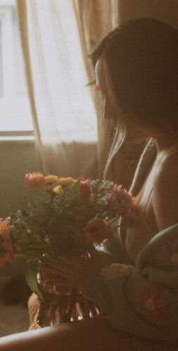 A person sits on a bed holding a bouquet of flowers. The room is softly lit with a window in the background.