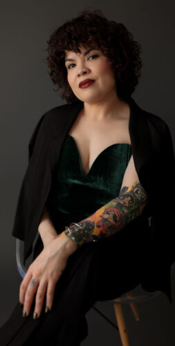 A person with curly hair sits confidently on a stool. They wear formal attire with a dark blazer over a green top and have a colorful tattoo sleeve on their left arm.