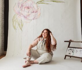 Woman sitting on the floor during a branding photoshoot, smiling, with a painted flower on the wall behind her.