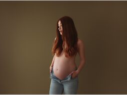 Pregnant person with long red hair participating in a maternity photoshoot, standing to the side and looking down at their belly against a brown background.