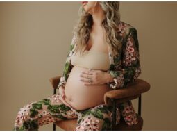 Pregnant woman in a Chelsea Football Club robe sitting on a chair, cradling her belly, with a neutral backdrop.