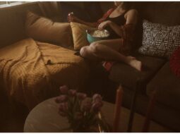 Kaitlyn relaxing on a couch with a bowl of popcorn, surrounded by pillows in a dimly lit room. There's a table with flowers in the foreground.