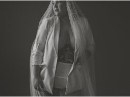 A black and white Denver Bridal Boudoir portrait of a bride in a veil and robe, revealing undergarments and a tattoo on her ribcage.
