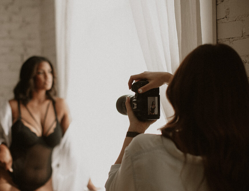 A photographer takes a picture of a woman posing in lingerie near a window with white curtains.