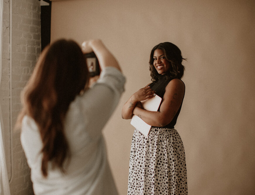 A woman holding a book poses near a beige wall while another person takes her photo in a room with natural light.