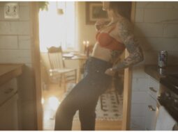 A woman in jeans and an orange crop top leans against a kitchen counter, sunlight streaming in, creating a warm, hazy glow.